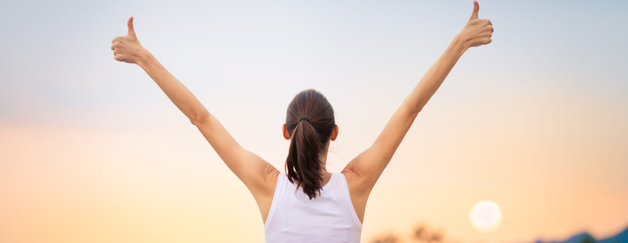 woman with arms raised in thumbs up facing a sunset